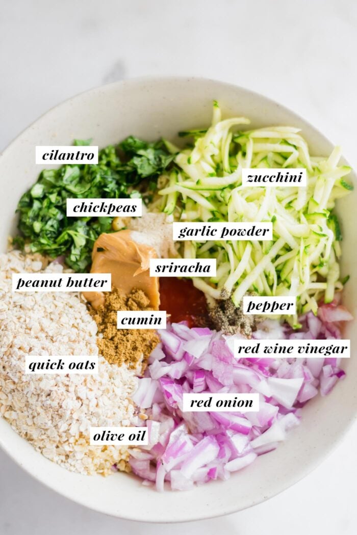Large mixing bowl with grated zucchini, cilantro, red onion, oats, peanut butter and spices. Each ingredient is labelled with text.