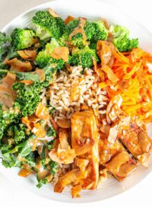 A bowl with tofu, sauce, kale, broccoli, carrot and coconut bacon.