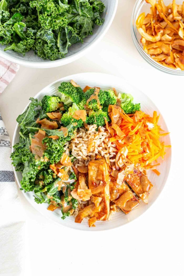 A bowl with tofu, sauce, kale, broccoli, carrot and coconut bacon.