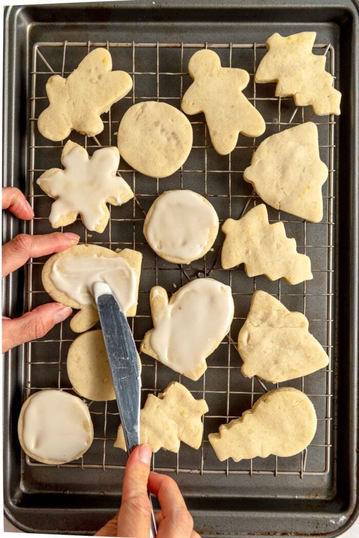 A hand using a knife to spread icing on a lemon shortbread cookie on a cooling rack.