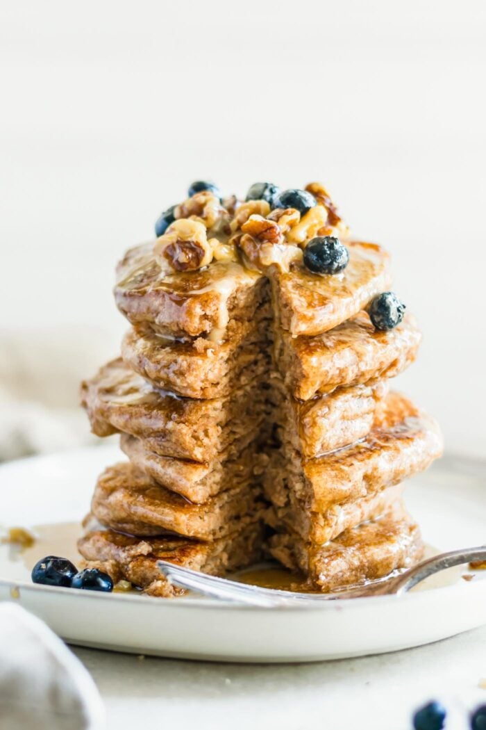 Stack of thick and fluffy whole grain pancakes with a slice taken from it. The pancakes are topped with blueberries, maple syrup and walnuts and a fork rests on the plate.