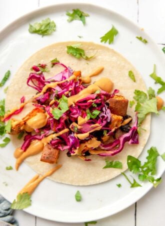 Overhead view of a colourful sweet potato and black bean taco topped with red cabbage slaw and a creamy sauce on a plate. Chopped fresh cilantro is sprinkled around the plate.