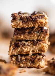 A stack of 4 seven layer cookies bars with a layer of graham cracker crust and a gooey layer of coconut, chocolate chips and pecans.