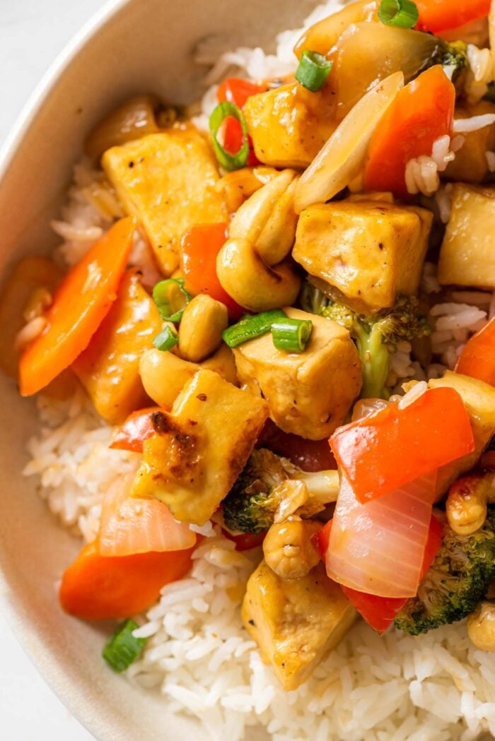Close up of a tofu stir fry with vegetables and cashews over rice in a bowl.