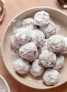 Overhead view of a bowl of powdered sugar-coated chocolate snowball cookies. A bowl of icing sugar can be seen just out of frame.