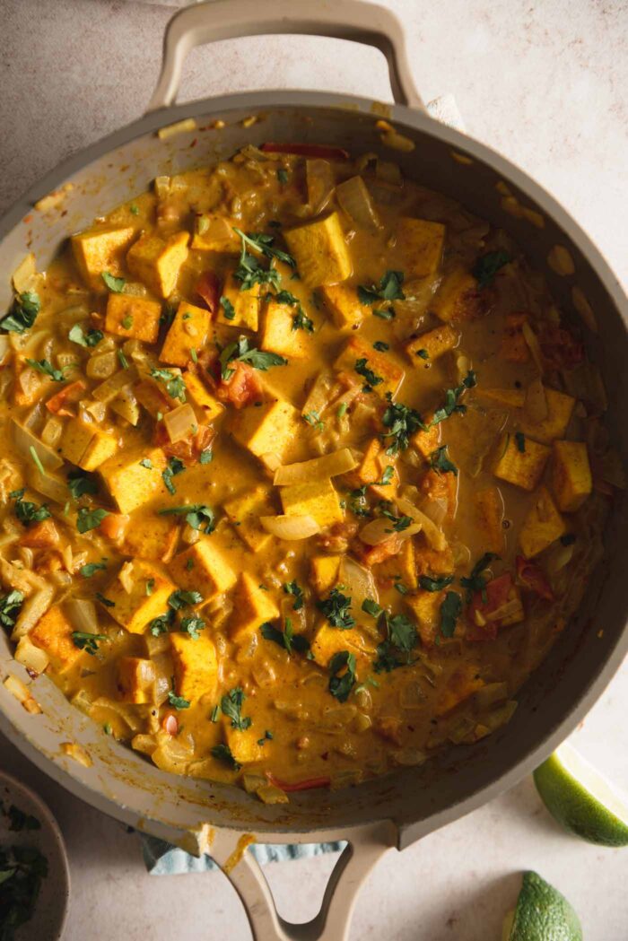 Cubes of tofu, diced onion and tomato cooking in a creamy coconut curry sauce in a skillet.