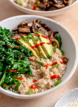 Bowl of savory oatmeal topped with avocado slices, cooked kale and mushroom and some hot sauce.