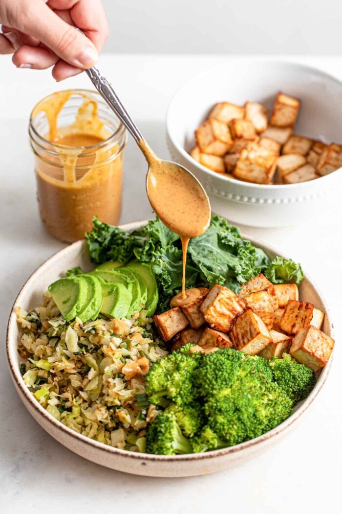 Drizzling a spoonful of sauce over a bowl with tofu, avocado and veggies.