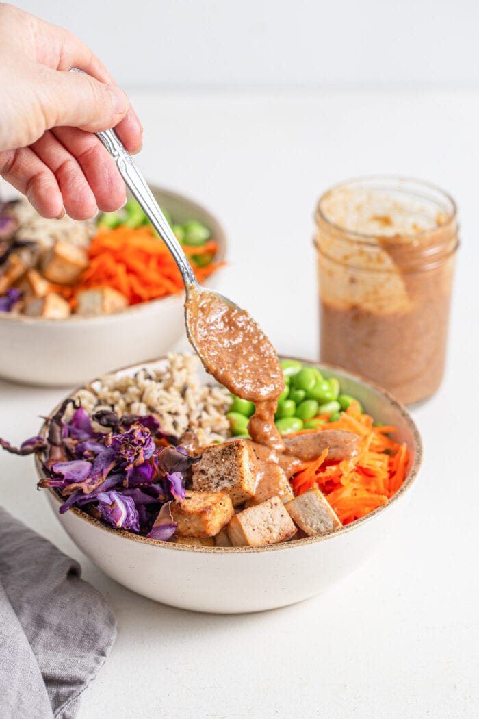 Hand spooning sauce over a colourful bowl of rice, tofu and vegetables.