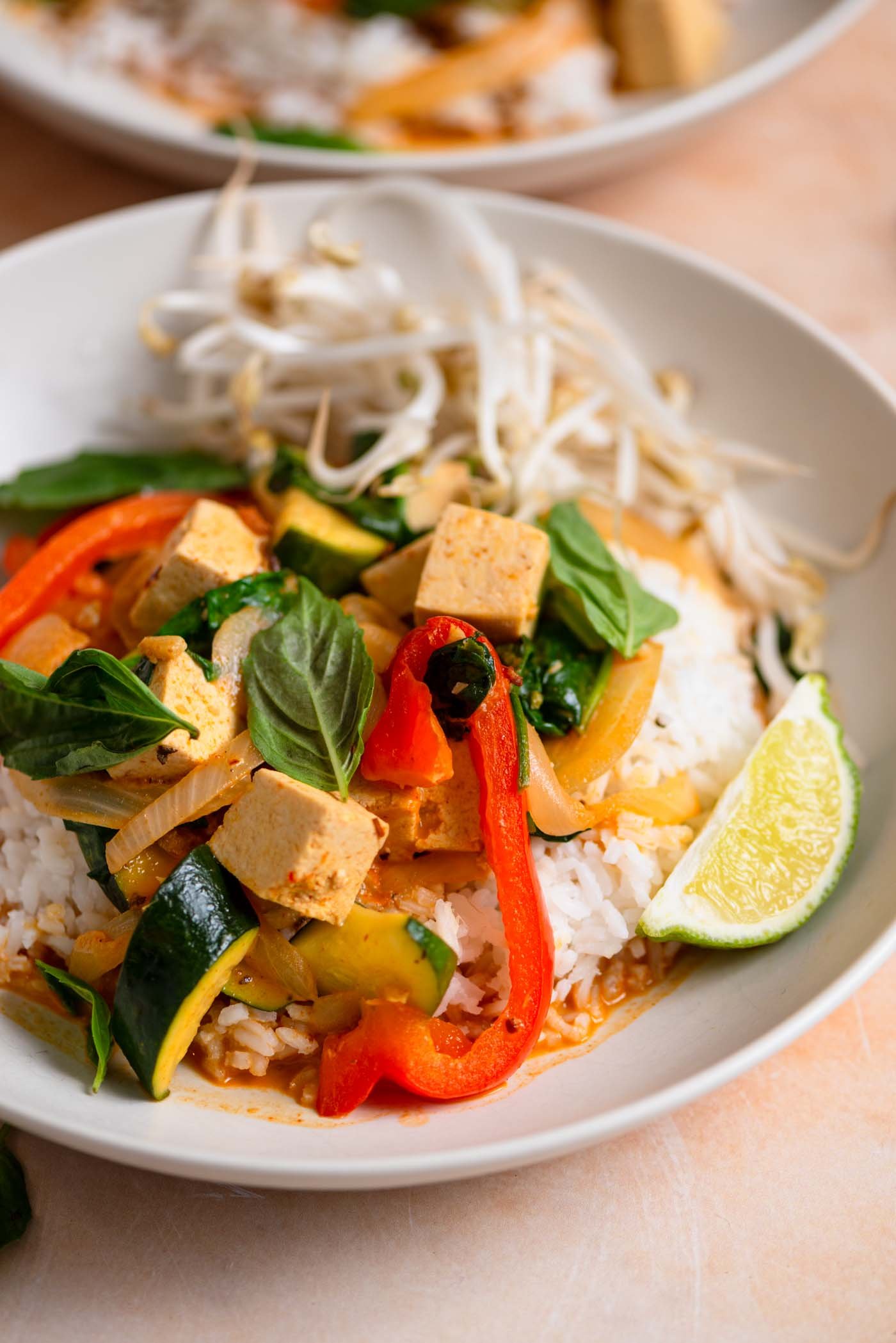 Bowl of Thai red curry with cubes of tofu, bell pepper and zucchini with fresh bean sprouts, basil leaves and a lime wedge.