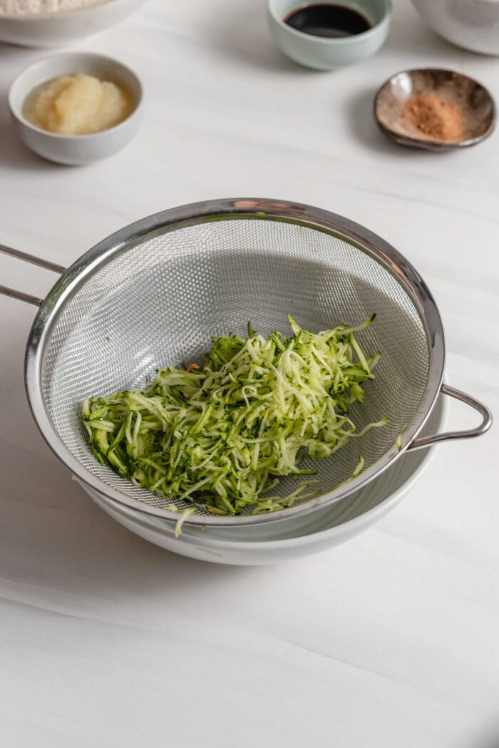 Grated zucchini in a strainer sitting in a bowl.